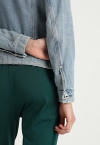 Jasnoniebieska kurtka dżinsowa o skróconym kroju, z widocznym przeszyciem i srebrnymi zapięciami na guzik na mankietach. Noszona na zielonych spodniach.