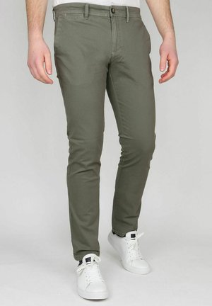Man die slim fit olijfgroene broek en witte sneakers draagt, staat tegen een effen achtergrond.