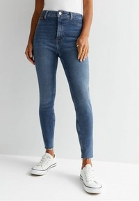 Höga, smala jeans i medelblå denim med subtila blekningar och fransade fållar, i kombination med vita sneakers.