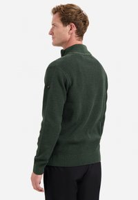 Man in een donkergroen, gestructureerd ritstrui gezien vanuit de achterkant, in combinatie met zwarte broek, staand tegen een witte achtergrond.