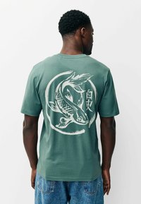 Zöld pamut póló, melynek hátoldalán egy nagy fehér koi hal mintája látható, körkörös ecsetvonás keretben. Rövid ujjú, laza szabású.