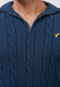 Bărbat îmbrăcat cu un pulover tricotat albastru, cu guler tip half-zip și un mic logo cu o pasăre galbenă pe partea stângă a pieptului.