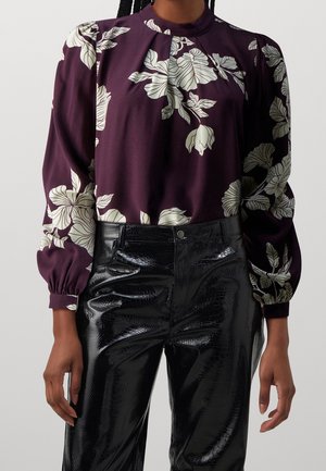 Mujer con blusa morada con grandes estampados florales blancos y pantalones negros de talle alto, brillantes y con textura.