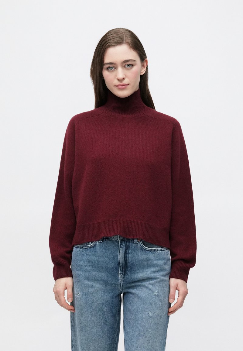 Bordowy golf o luźnym kroju z prążkowanymi mankietami i dołem, zestawiony ze światłoniebieskimi, postrzępionymi dżinsami. Miękka faktura, design sięgający do połowy tułowia.