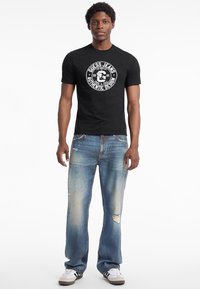 Sort t-shirt med et cirkulært grafisk logo, parret med slidte blå jeans. Modellen har hvide sneakers med sorte striber på.