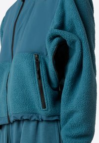 Teal fleecová bunda s hladkým svrchním materiálem, bočními kapsami na zip a texturovaným, nadýchaným materiálem na rukávech a těle.