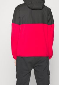 Chaqueta con capucha que presenta una sección superior negra y una sección inferior roja. Fabricada con tela suave, tiene puños elásticos y dobladillo ajustable.