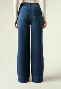 Ciemnoniebieskie dżinsy typu wide-leg, wyróżniające się wysokim stanem, tylnymi kieszeniami i prostym wykończeniem. Miękka tekstura denimu o gładkim wykończeniu.