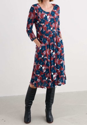 Frau trägt ein knielanges, langärmeliges rotes Kleid mit blauem und weißem Blumenmuster und schwarze kniehohe Stiefel, Hände in den Taschen.