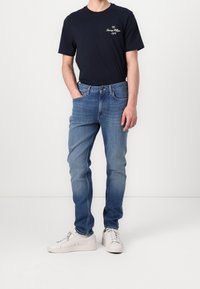 Hombre vestido con una camiseta de color azul marino oscuro con el logo de Tommy Hilfiger, unos pantalones vaqueros ajustados de color azul claro y zapatillas blancas, de pie con una mano en el bolsillo.