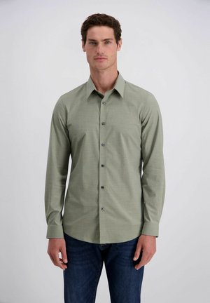 Mann trägt ein hellgrünes, langärmliges Hemd mit Knöpfen und dunkelblaue Jeans und steht vor einem schlichten weißen Hintergrund.