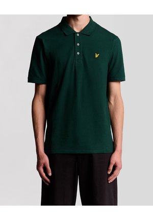 Homme portant un polo vert foncé à manches courtes avec trois boutons et un petit logo d'oiseau jaune sur la poitrine gauche.