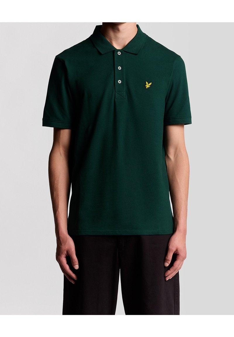 Homme portant un polo vert foncé à manches courtes avec trois boutons et un petit logo d'oiseau jaune sur la poitrine gauche.