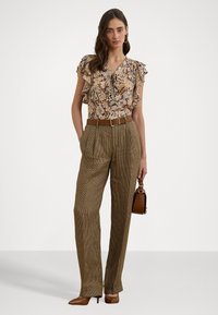 Bluse mit floralen Mustern und Rüscheln, kombiniert mit beigefarbenen, karierter, hoch taillierter Hose, braunem Gürtel und passender Handtasche. Braune Schuhe mit Absatz.