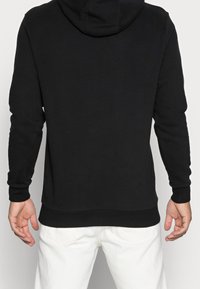 Sweat à capuche noir à la texture douce, avec des manches longues, une grande capuche, et une ceinture ainsi que des poignets en bord-côtes. Porté avec un pantalon blanc.