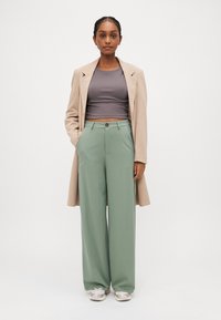 Beige Mantel, graues gekürztes Oberteil und mintgrüne weit geschnittene Hosen. Turnschuhe in Mischfarben. Modell steht mit Händen in den Taschen.