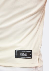 Gros plan de l'ourlet d'un t-shirt de sport couleur crème avec une étiquette noire indiquant "Engineered to the exact specifications of championship athletes."