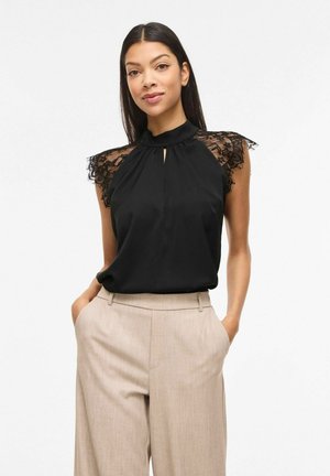 Kobieta w czarnej, bezrękawnikowej bluzce z koronkowymi ramionami i beżowych, szerokich, wysokich spódnicach, z rękami w kieszeniach, na jednolitym tle.