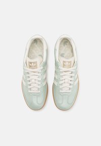 Une paire de baskets Adidas vert clair avec des rayures blanches, des patchs de logo beige, des lacets blancs et des semelles en gomme, présentée de dessus sur un fond blanc.