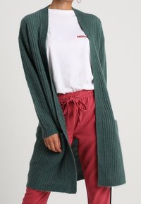 Cardigan lungo con apertura frontale in maglia a costine color verde scuro. Caratterizzato da maniche lunghe, due tasche laterali, indossato sopra una t-shirt bianca e pantaloni rosa.