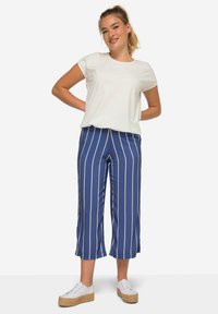 Bílý top s krátkým rukávem, modré pruhované kalhoty s širokými nohavicemi a krémovými pruhy, a bílé tenisky s jutovými podrážkami. Hladká textura látky.