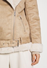 Beige jacka i konstmocka med en krage i fårskinn, dragkedja, sidofickor och ett skärp runt midjan med en silverfärgad spänne.