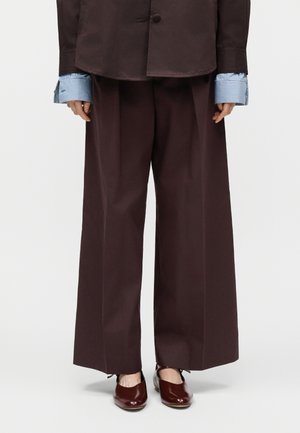 Pantalones anchos de color marrón oscuro combinados con una chaqueta a juego, puños de camisa a rayas azules visibles y zapatos planos marrón oscuro brillantes.