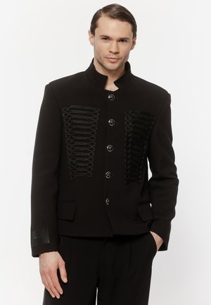 Mężczyzna w czarnej kurtce z wysokim kołnierzem, ozdobionej pętlami i srebrnymi guzikami, stoi z jedną ręką w kieszeni na białym tle.