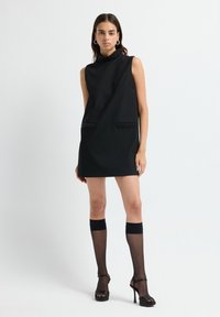 Frau trägt ein ärmelloses schwarzes Mini-Kleid mit Perlenkragen und Taschen, durchsichtige knielange Socken und schwarze High Heels vor weißem Hintergrund.