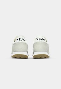 Hvite og grå sneakers med en teksturert semsket hælkappe, svart "VEJA"-merke, og en formstøpt gummisåle med et grep-mønster.