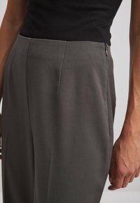 Szare wykończone spodnie o gładkiej fakturze, z przodami z zakładkami i bocznym zamkiem błyskawicznym. Zbliżenie na pasek i ułożenie rąk.