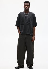 T-shirt nera oversize con dettaglio testurizzato "89", abbinata a pantaloni larghi verde scuro e scarpe nere lucide. Accenti in catena argentata visibili.