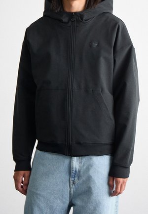 Osoba ubrana w czarną bluzę z zamkiem błyskawicznym z przodu i kieszeniami oraz jasnoniebieskie dżinsy, pokazana od ramion do bioder na jednolitym tle.