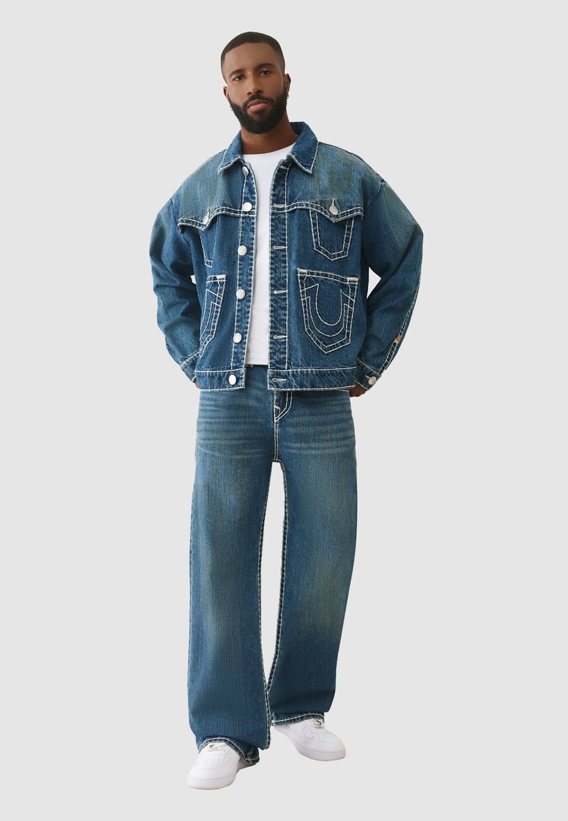 Veste en jean et jeans à jambes larges de couleur bleu moyen. La veste présente de grandes poches et des coutures contrastantes. Le look est complété par un t-shirt blanc et des baskets.