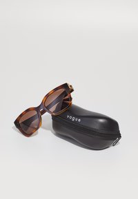 Ochelari de soare din scoică cu lentile maro, formă pătrată, și accent de aur lângă braț, așezați lângă un toc negru cu fermoar, etichetat "vogue."