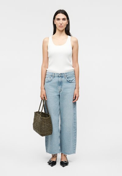 Γυναίκα με μακριά σκούρα μαλλιά που φοράει λευκό τοπ, ανοιχτό μπλε τζιν με φαρδιά πόδια, μαύρα μυτερά παπούτσια και κρατάει πλεκτή καφέ τσάντα.