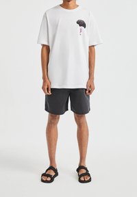 T-shirt en coton blanc à manches courtes, avec un motif graphique noir et un texte rose. Porté avec un short foncé et des sandales noires.