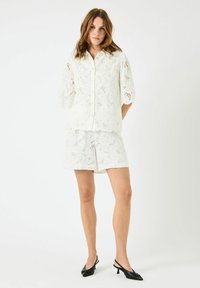 Hvit, brodert bluse med knapping og korte, puffede ermer, sammen med matchende shorts. Svarte slingback hæler fullfører looket.