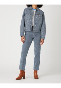 Kvinna iklädd en blårandig och vit denimjacka och byxor med vit skjorta och bruna klackiga ankelstövlar, stående.