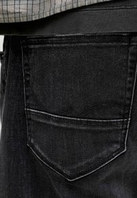 Gesäßtasche einer schwarzen Jeans, getragen mit einem hineingesteckten grau-weiß karierten Hemd.