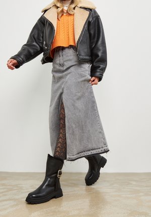 Osoba v černé koženkové bundě, oranžovém zkráceném svetru, dlouhé šedé džínové sukni s bočním rozparkem, vzorovaných punčochách a černých botách.