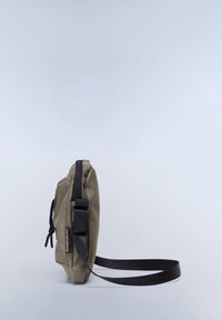 Olive green canvas crossbody bag with a black strap, zip closure, and branded patch. Compact, rectangular shape with textured fabric.