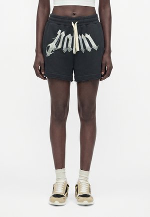 Person iført sorte shorts med hvid snor og stiliseret tekst, hvide crew-sokker og beige og sorte sneakers, stående mod en hvid baggrund.