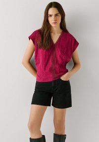 Vrouw met lang bruin haar, gekleed in een rode blouse met V-hals en geborduurde rand, zwarte denim shorts en zwarte laarzen tot halverwege de kuit, staand tegen een witte muur.