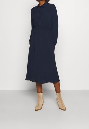 Personne portant une robe midi bleu marine à manches longues avec une patte de boutonnage à l’avant et des bottines beiges, debout devant un fond blanc uni.