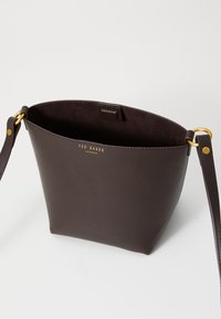 Sac seau en cuir marron avec un logo doré, base ronde, doublure intérieure et une bandoulière amovible ; texture lisse, design minimaliste.