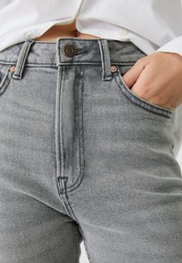 Jeans de denim gris con una cintura alta, costuras visibles en los bolsillos frontales y detalles de hardware en bronce. La tela tiene una textura sutil.