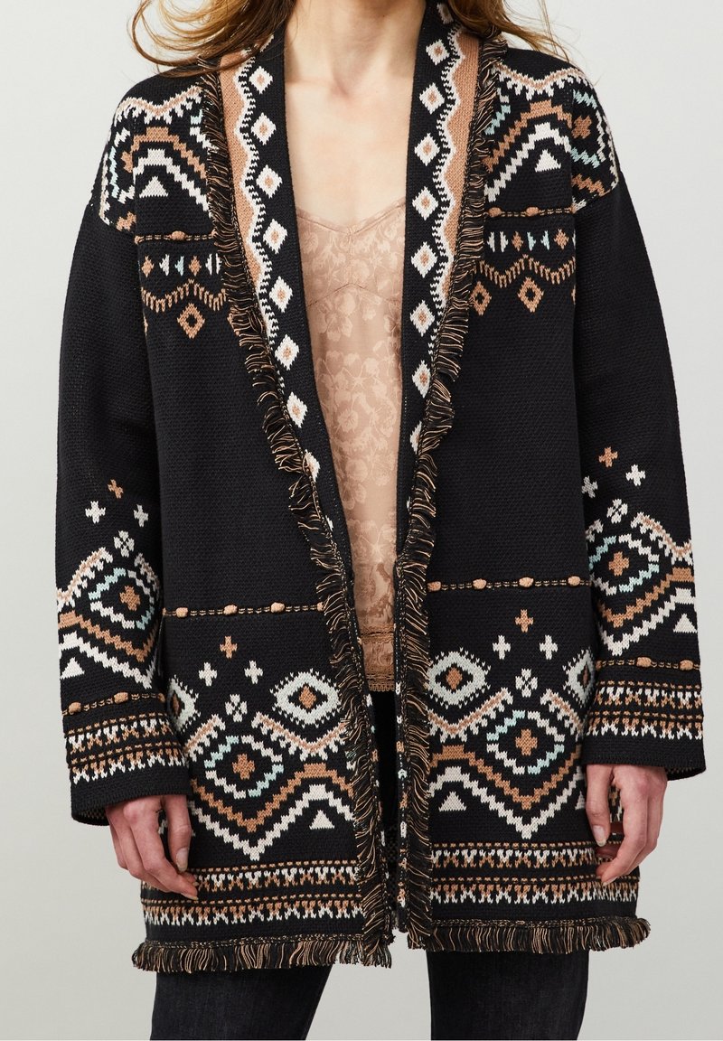Femme portant un cardigan noir avec des motifs géométriques marron, blanc et bleu sarcelle et des bords à franges par-dessus un haut en dentelle beige.