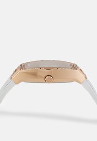 Montre rectangulaire au cadran doré, avec un bracelet blanc, dotée d'une lunette texturée et d'une couronne sur le côté, surface en verre lisse et design minimaliste.