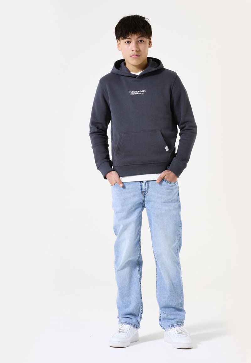 Sudadera gris oscuro con bolsillo frontal, con texto blanco "OBJETIVOS FUTUROS", combinada con jeans azul claro y zapatillas blancas. Tela suave, corte casual.